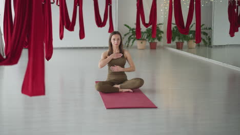 yoga instructor seated on red mat places hand on chest and other on stomach while focusing on breath and posture amid aerial studio setting with hanging red silk hammocks and mirrored backdrop