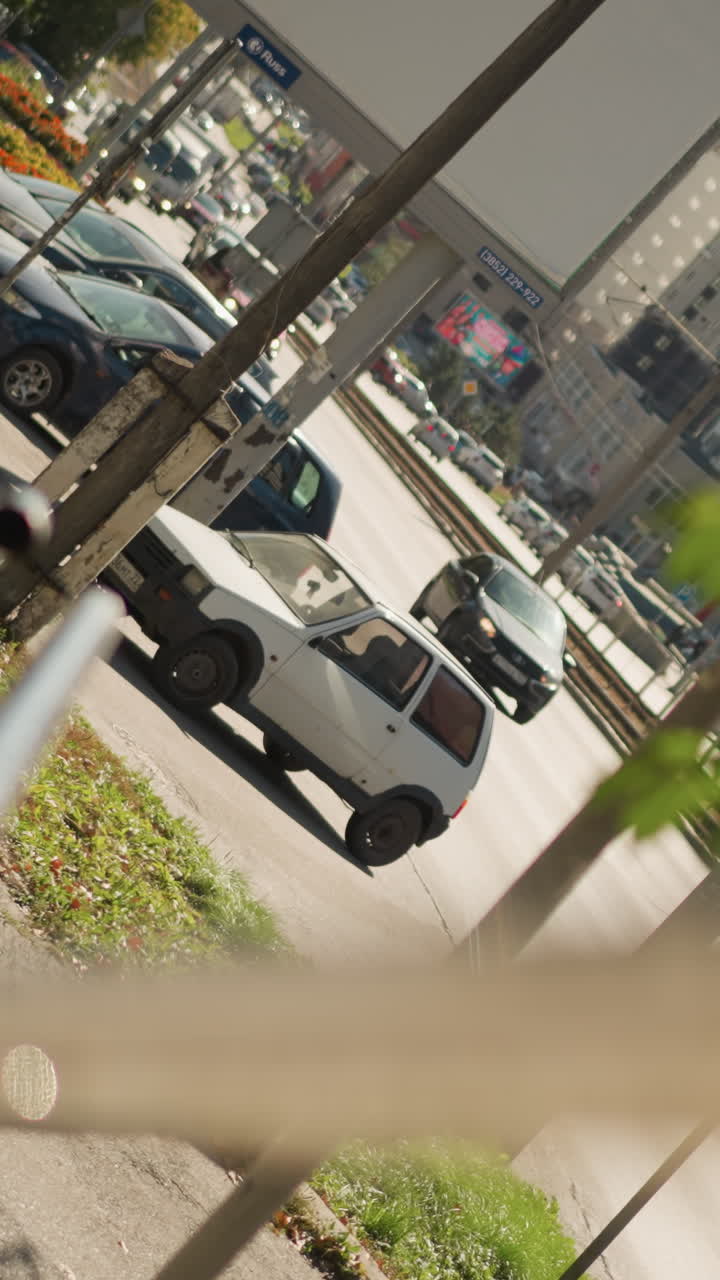 붐비는 교차로에서 회전하는 흰색 자동차, 흐릿하게 보이는 난간이 보도와 쉼터를 액자처럼 감싸고 있으며, 여름 햇빛 아래 창문이 반사되는 가운데 주차된 차량과 이동하는 차량 행렬, 솔직한 도시 풍경