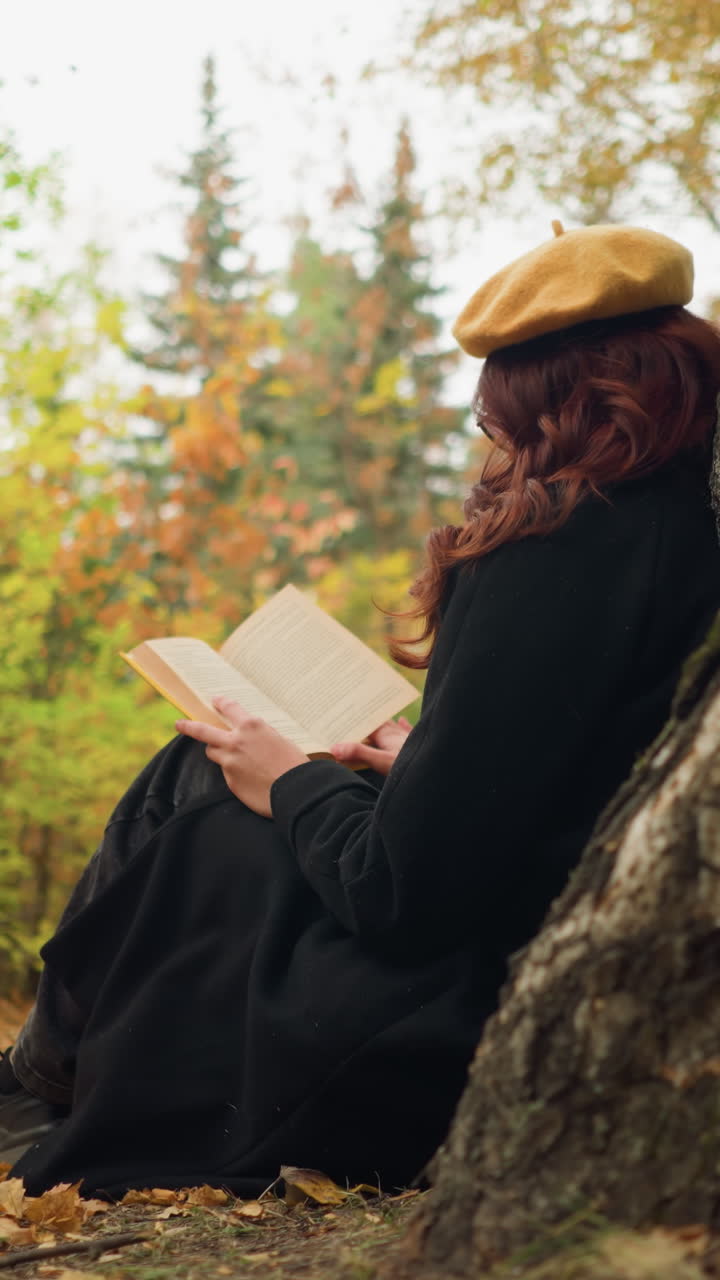 mujer concentrada sentada al aire libre en el bosque de otoño apoyada en un árbol, profundamente absorta en el libro, con boina amarilla y abrigo negro, disfruta de la tranquila soledad rodeada de vibrante follaje de otoño