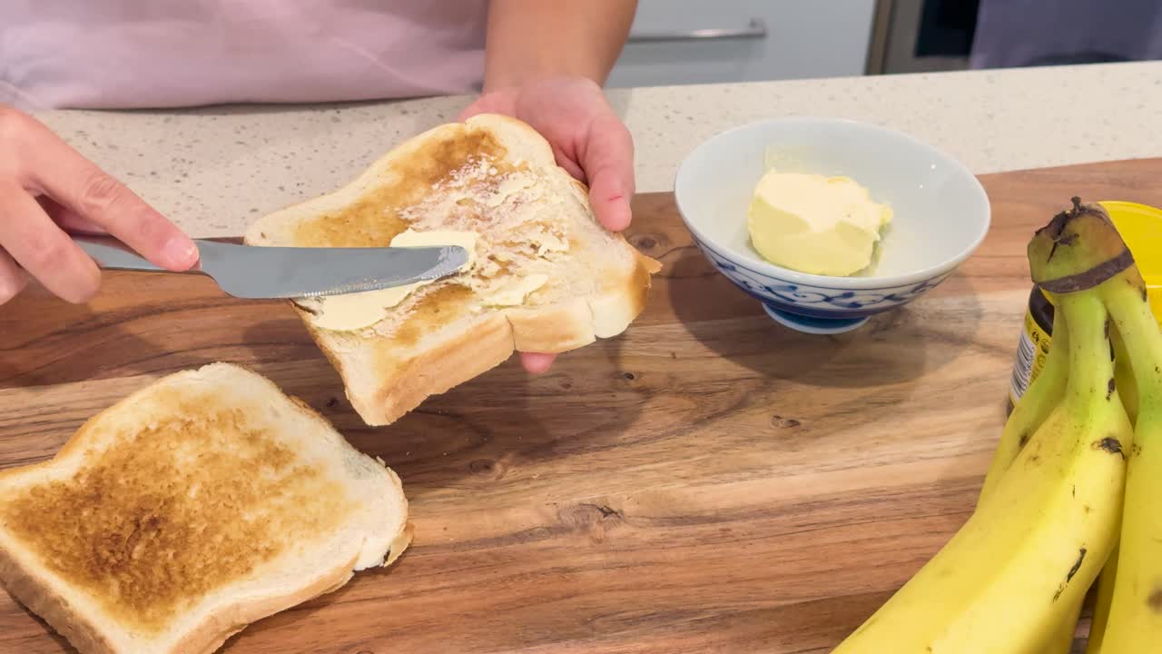 A person spreads butter onto slices of toasted bread on a wooden countertop, with bananas and a butter dish visible, under bright indoor lighting