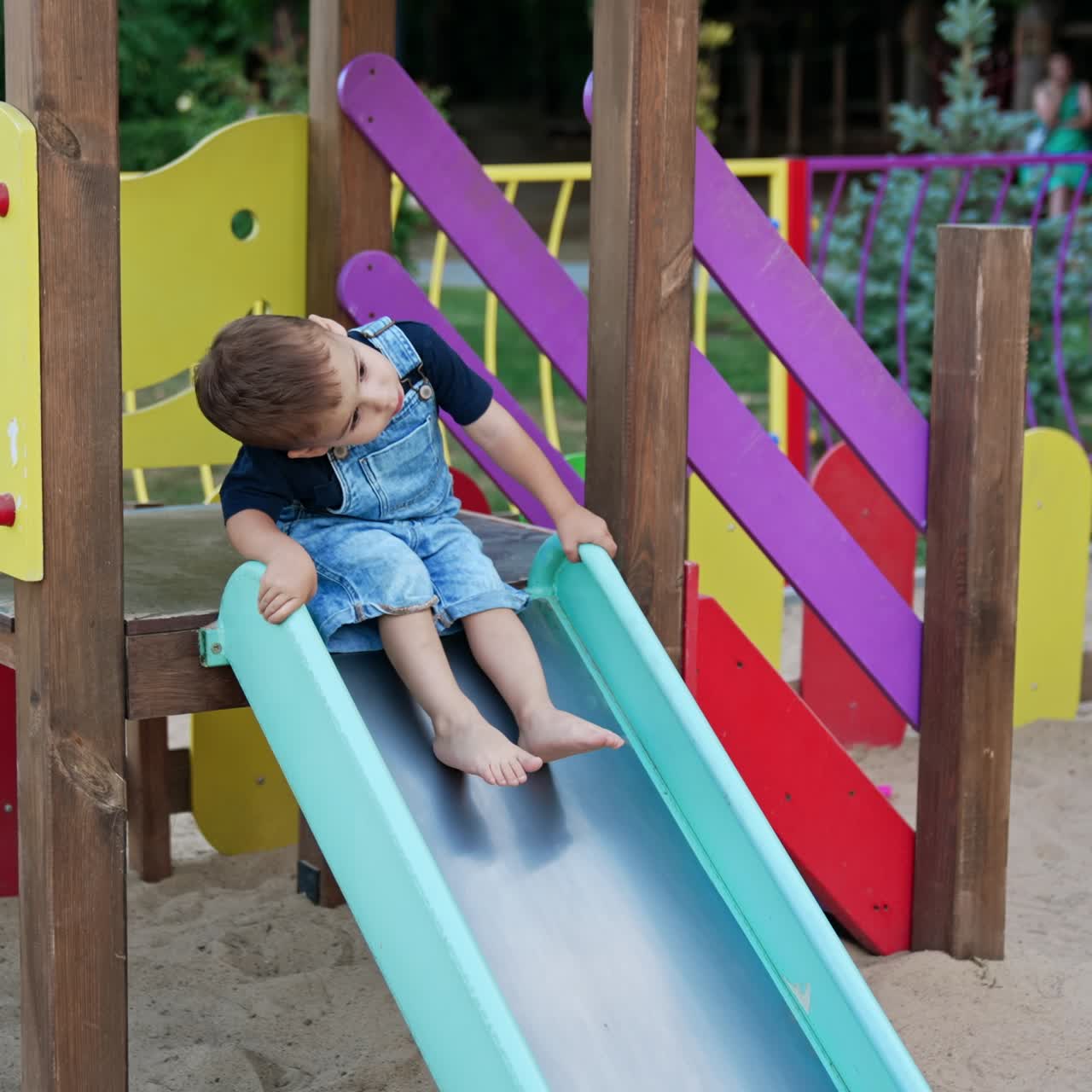 Cute toddler in a jeans romper sitting on the slide. Kid bends to the side playing on the playground in summer