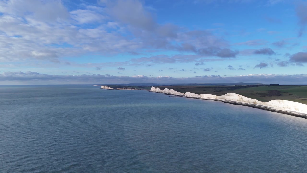 Doğu Sussex, İngiltere, Birleşik Krallık'ta kıyı şeridi boyunca uzanan Seven Sisters beyaz tebeşir kayalıklarının, sakin mavi deniz ve geniş bulutlu gökyüzü ile havadan drone görüntüsü.