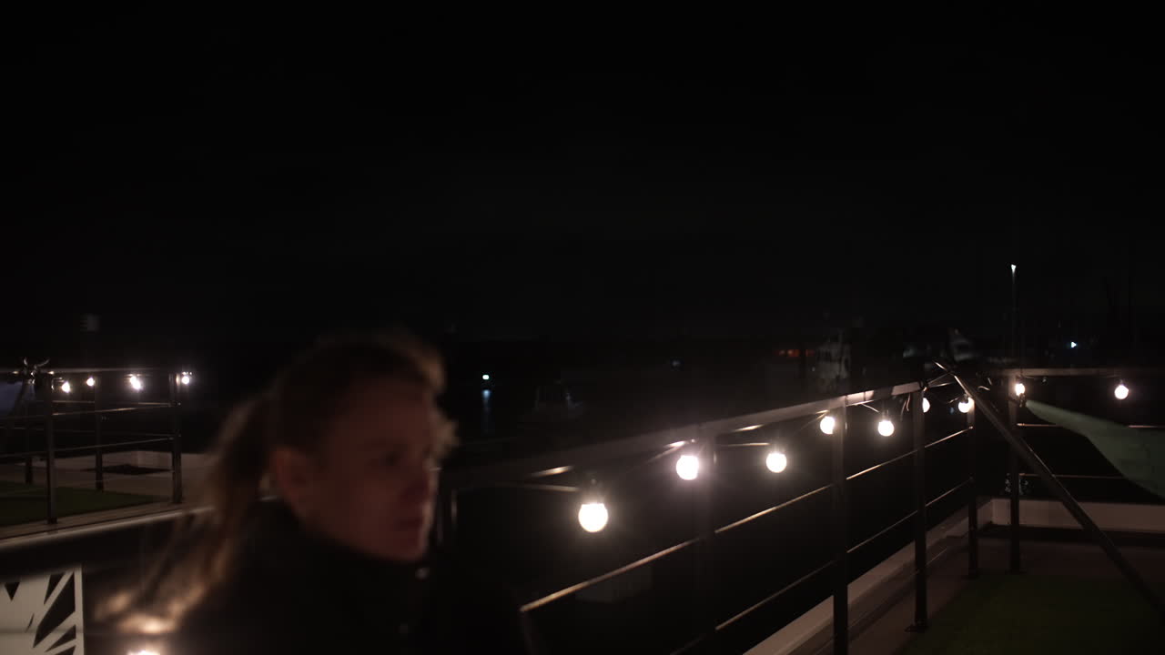Young woman enjoying cold beer during nighttime terrace gathering, surrounded by soft string lights creating intimate urban atmosphere and relaxed mood. Her face is in blur, filmed with vintage lens