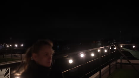 young woman enjoying cold beer during nighttime terrace gathering, surrounded by soft string lights creating intimate urban atmosphere and relaxed mood. her face is in blur, filmed with vintage lens