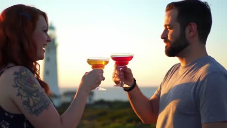 a romantic sunset toast: capturing the joyful moments of two friends celebrating with colorful cocktails near a glimmering lighthouse on the coast