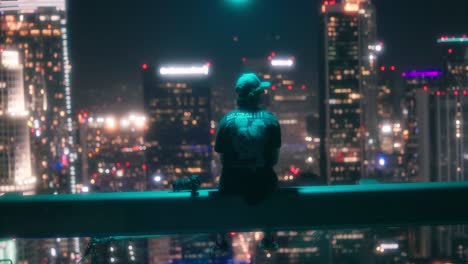 adulto joven sentado en la cornisa de un rascacielos mira hacia el centro de los ángeles por la noche