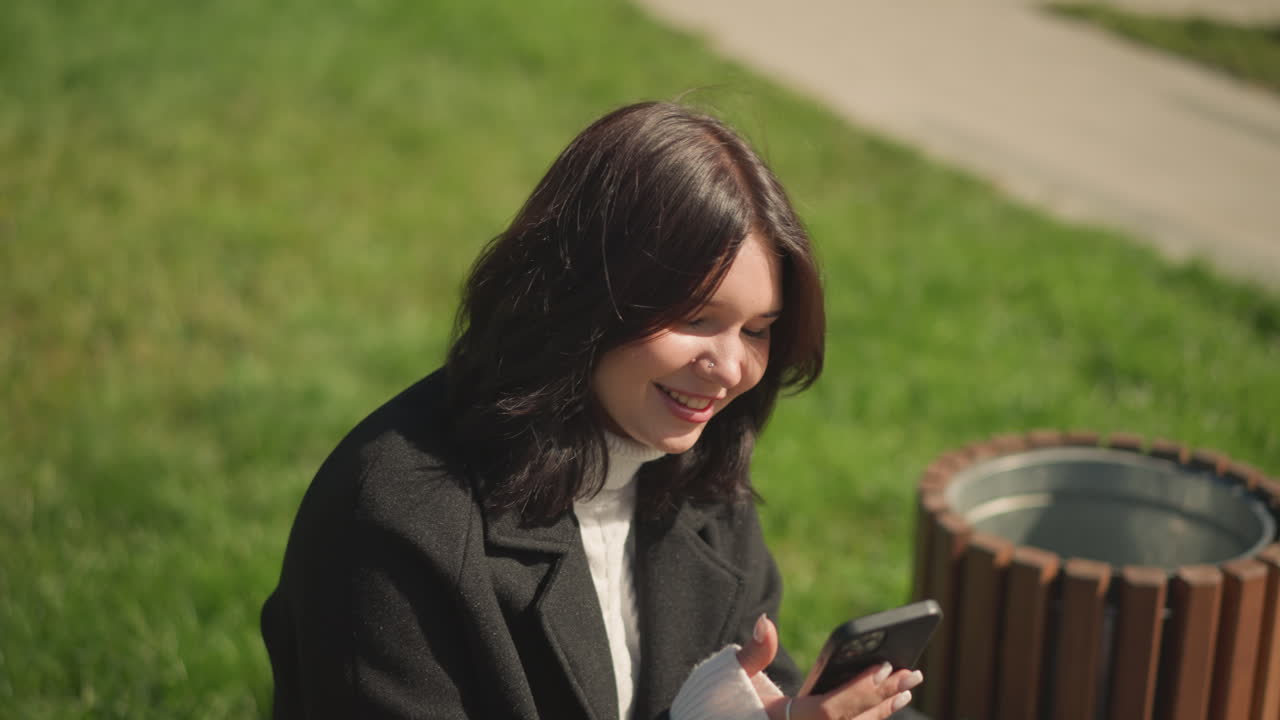 쾌활한 여성이 웃고 머리카락을 조정하면서 드폰에 초점을 맞추고, 배경에 초록색이 있고, 편안한 야외 공원 분위기입니다.