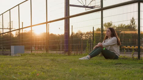 lonely woman sits on lawn of sports stadium at sunset. last sun rays illuminate sad lady thinking about going to psychologist to solve problems. female rests alone