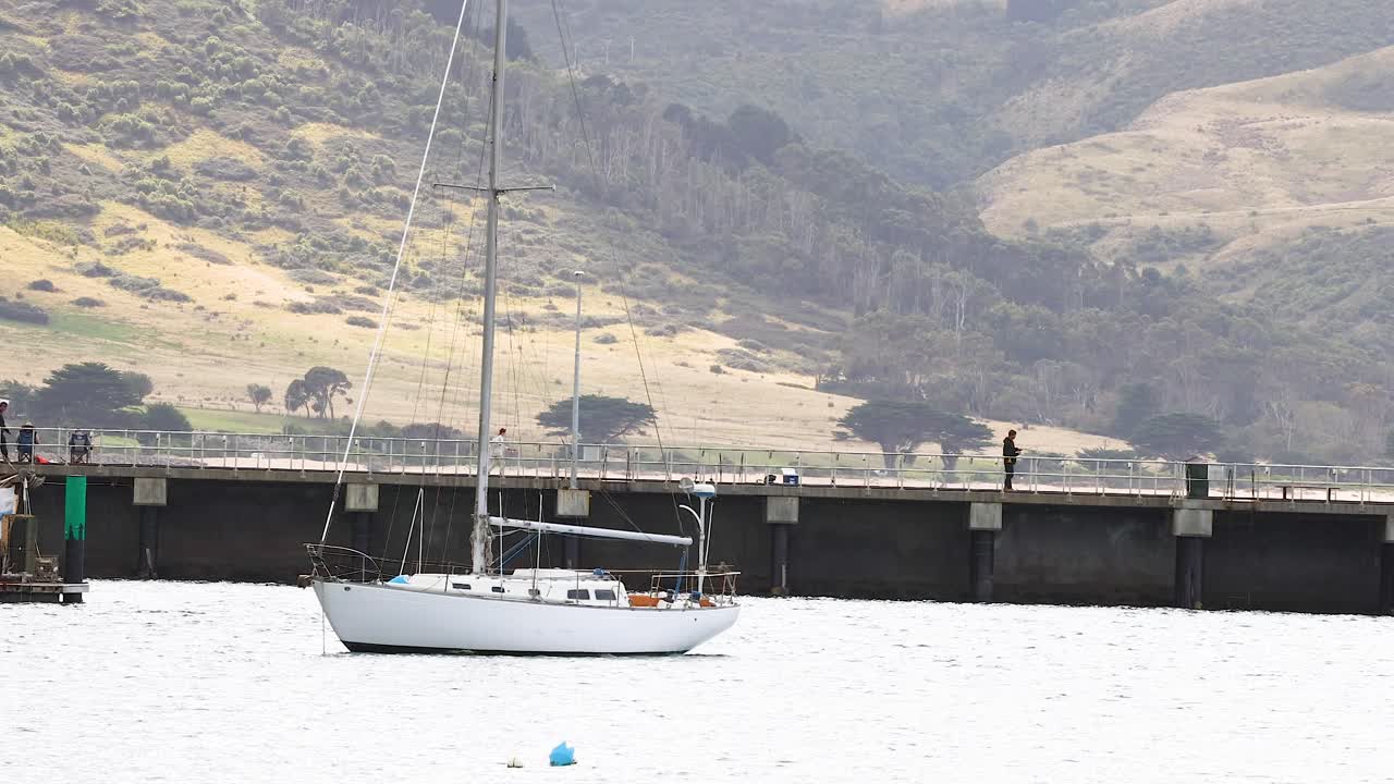 A sailboat is stationary near a pier with scenic hills in the background. The lighting is soft, creating a serene atmosphere