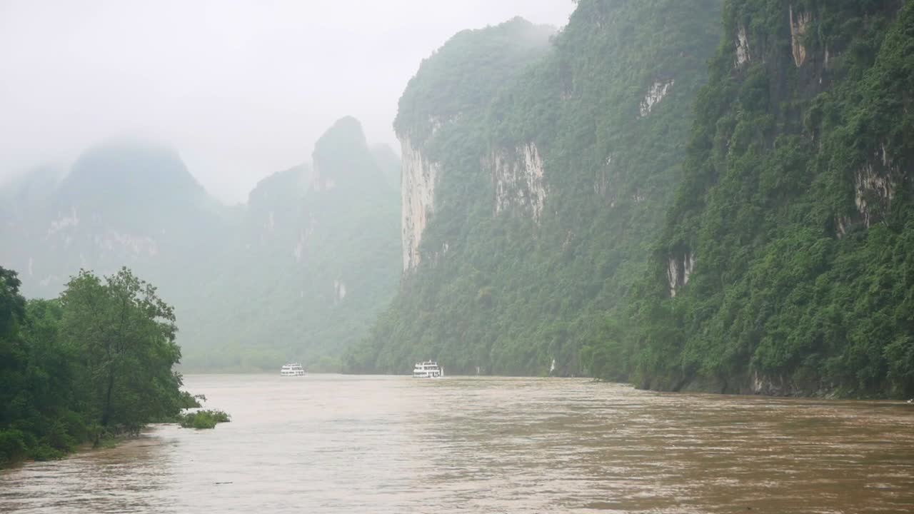 Sejltur på en båd langs en brun flod i Guilin, Kina. Betragter de tågede grønne bjerge i baggrunden.