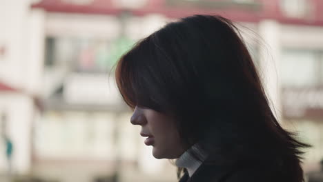 woman with soft brown hair and contemplative expression captured in side profile in outdoor setting, surrounded by blurred urban background and soft natural light