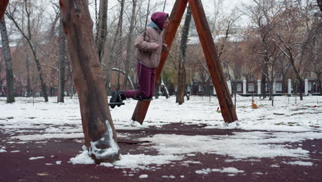 side view of lady dressed for winter in maroon trousers on swing holding chains with snow on the ground and bare trees in the background, enjoying outdoor activity in cold weather