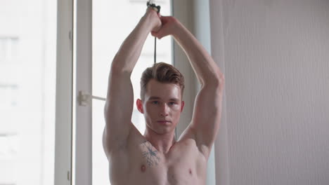 determined student standing near window with arms raised pulling resistance band overhead, focused expression on face, natural sunlight filtering through window