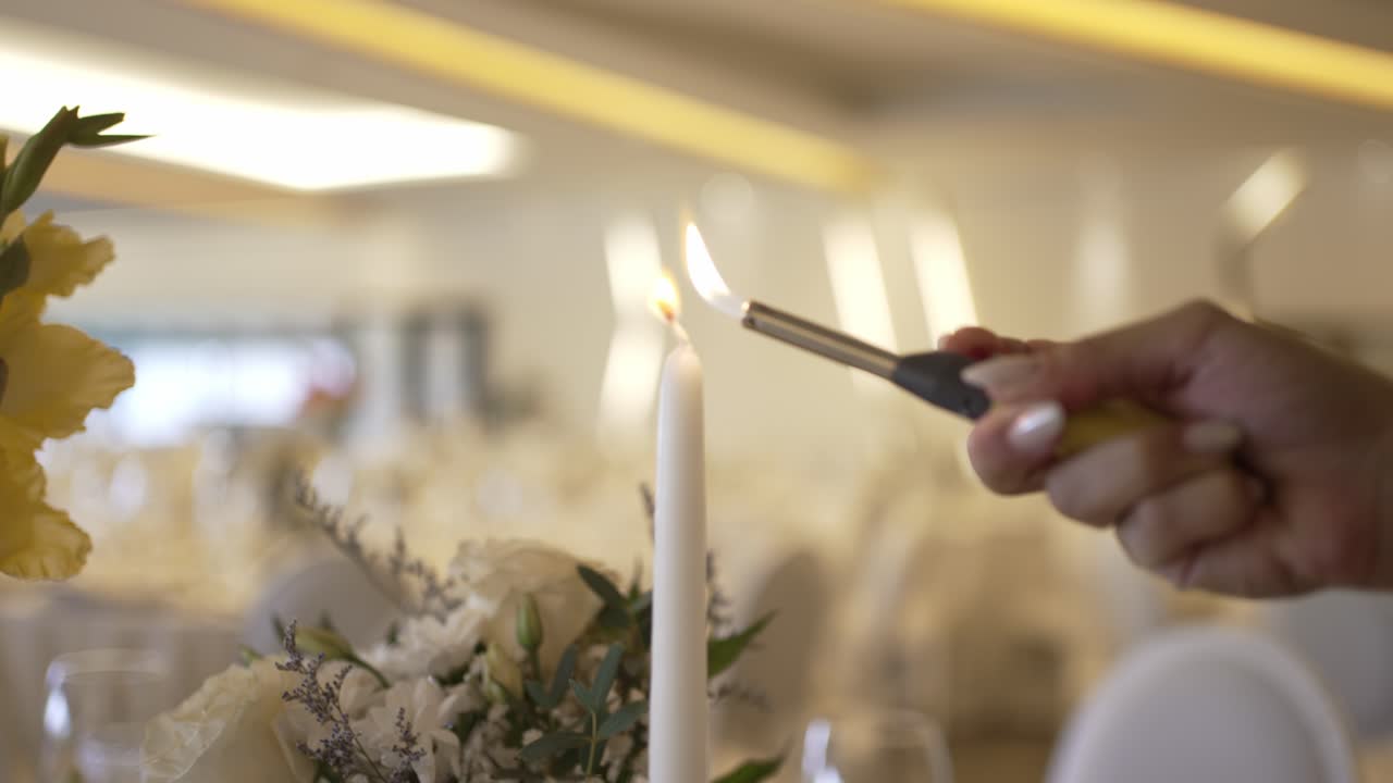 mano usando un encendedor para encender una vela cónica blanca, puesta contra un fondo de salón de banquete de enfoque suave