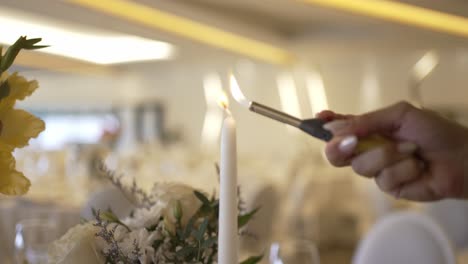 hand using a lighter to light a white taper candle, set against a soft focus banquet hall backdrop