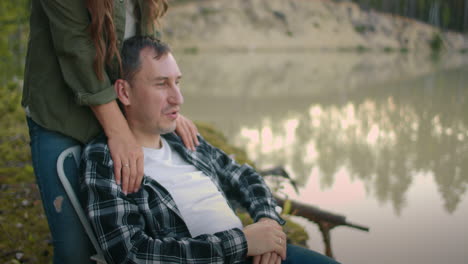 loving spouses are resting at nature man is sitting in armchair on shore of calm lake in forest woman is stroking his shoulders