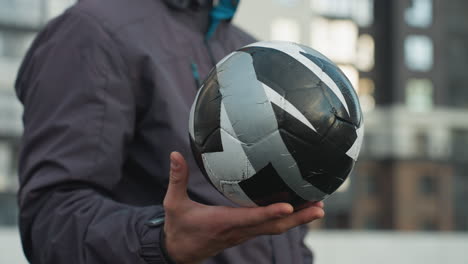 hombre hábilmente girando una pelota de fútbol en la palma de la mano con expresión enfocada, fondo borroso con edificios urbanos de gran altura y barra de malla