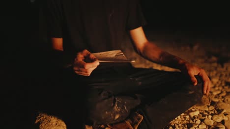 man sitting on ground in glow of burning fire picks up torn out diary entry