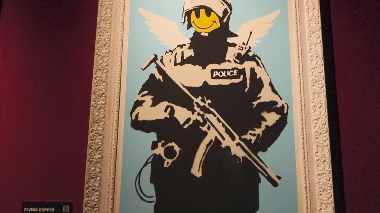 Multi-layered stencil graffiti painting artwork by Banksy called Flying copper at an exhibition. Shows a police officer standing with a weapon on blue background and a yellow smiley face on his face.