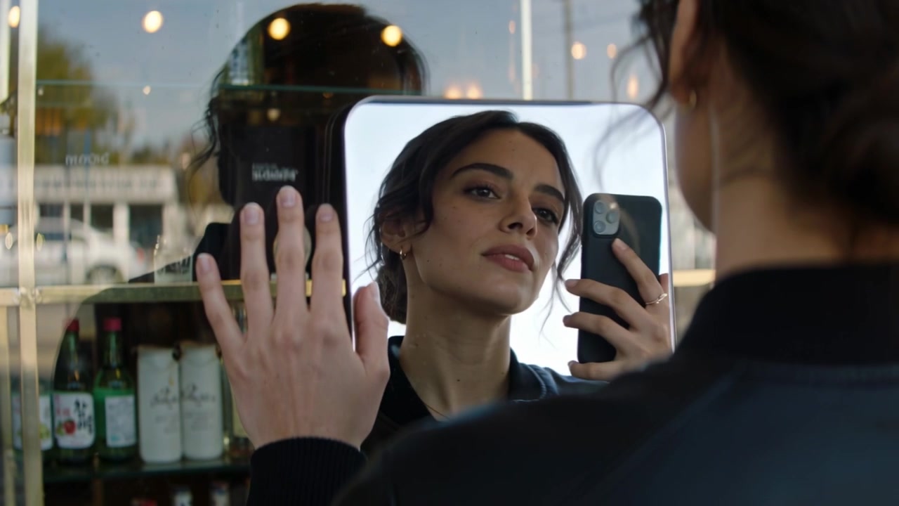 Donna con giacca nera che preme la mano su uno specchio in un negozio, scattando una foto con il telefono, con bottiglie sullo sfondo.