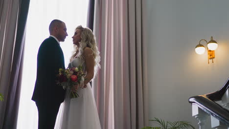 Loving-bride-with-bouquet-and-groom-stand-by-window-on-stairs