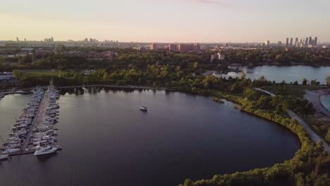 aerial sunset descending shot of sailboat and marina yacht club dock in lake bay surrounded by green trees with city buildings skyline in background in toronto ontario canada