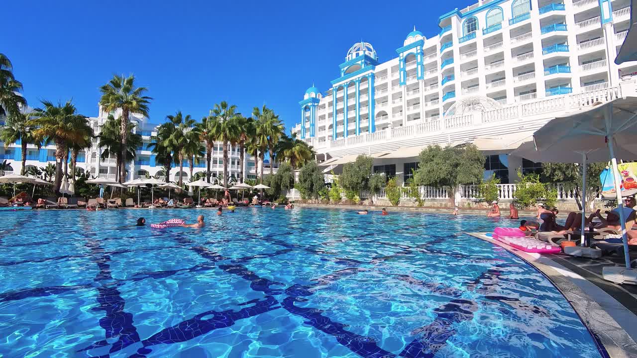 Luxury resort with a large outdoor swimming pool, surrounded by palm trees and sun loungers, with a bright blue sky and elegant white building in the background, perfect for vacation and relaxation.
