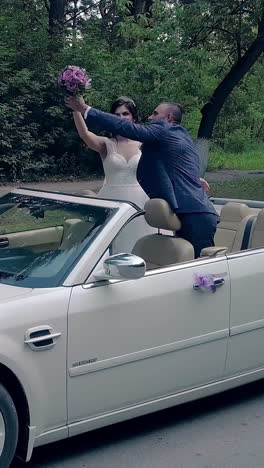 handsome fiance kisses pretty bride in white dress holding purple bouquet in celebratory convertible