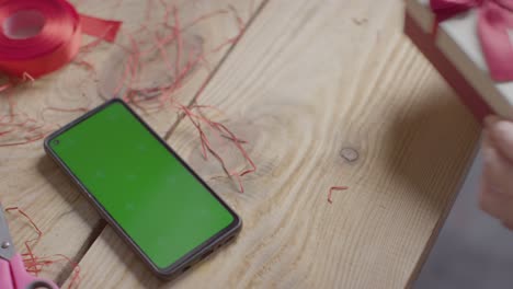 close up shot of man gift wrapping romantic valentines present in box next to green screen mobile phone