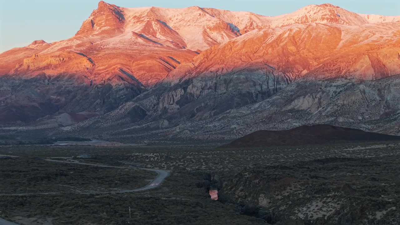 Un video panoramico che cattura la luce dorata dell'alba che illumina le cime montuose vulcaniche spolverate di neve sopra una vasta valle desertica nella Riserva La Payunia