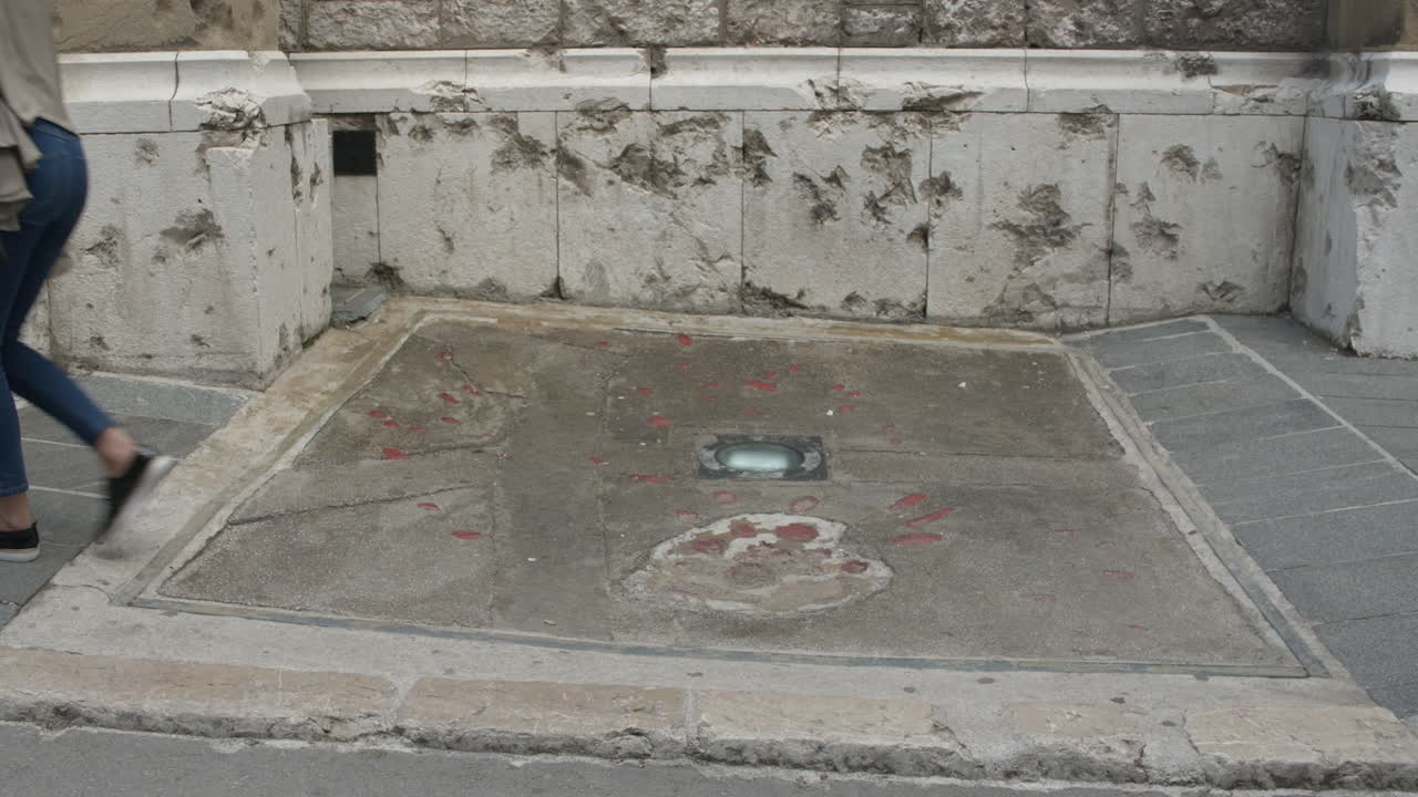 Pedestrians walk in foreground of war shelling damage and Sarajevo rose exterior of chapel. War memorial