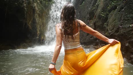 A-young-woman-with-long,-dark-hair-is-walking-with-her-yellow-pants---skirt-into-the-water-towards-a-small-waterfall