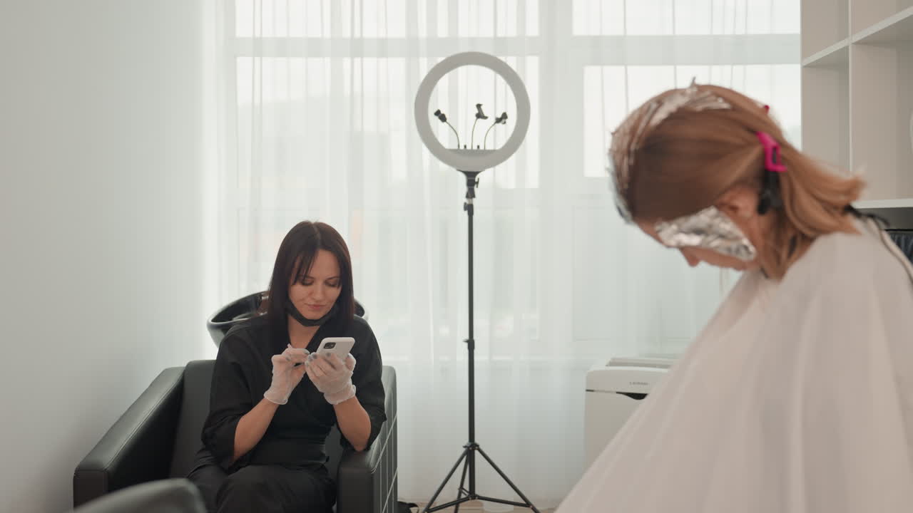 Cliente che scorre sullo smartphone in un salone, parrucchiere che applica la carta stagnola sui capelli, cliente seduto su una sedia nera che indossa un mantello, ring light vicino alla finestra, luce diurna soffusa che illumina l'interno minimal, arredamento moderno