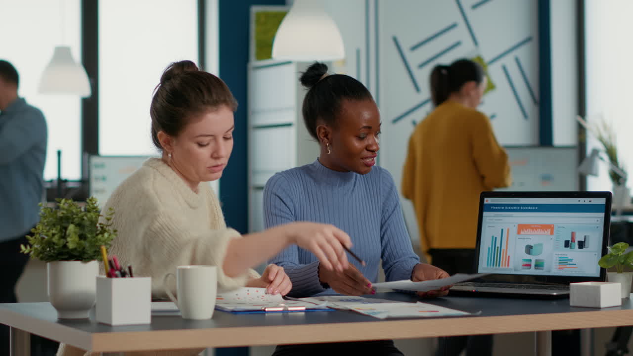 Startup employee working with african american coworker on group project comparing sales charts on clipboard