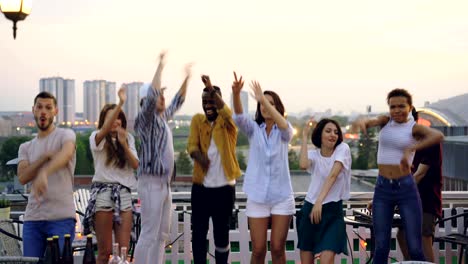 Sad people are standing on rooftop and looking at camera then starting dancing and jumping having fun with happy faces while DJ is working with mixing console.