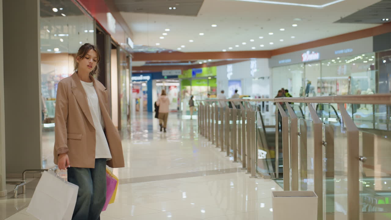 젊은 백인 브이로그 운영자가 쇼핑몰에서 쇼핑백을 열고, 구매한 물건을 공개하면서 호기심 가득한 표정을 짓고 있어요. 다채로운 쇼핑백과 부티크 인테리어가 장난기 넘치는 언박싱 장면을 연출하며, 라이프스타일 콘텐츠 분위기를 자아내요.