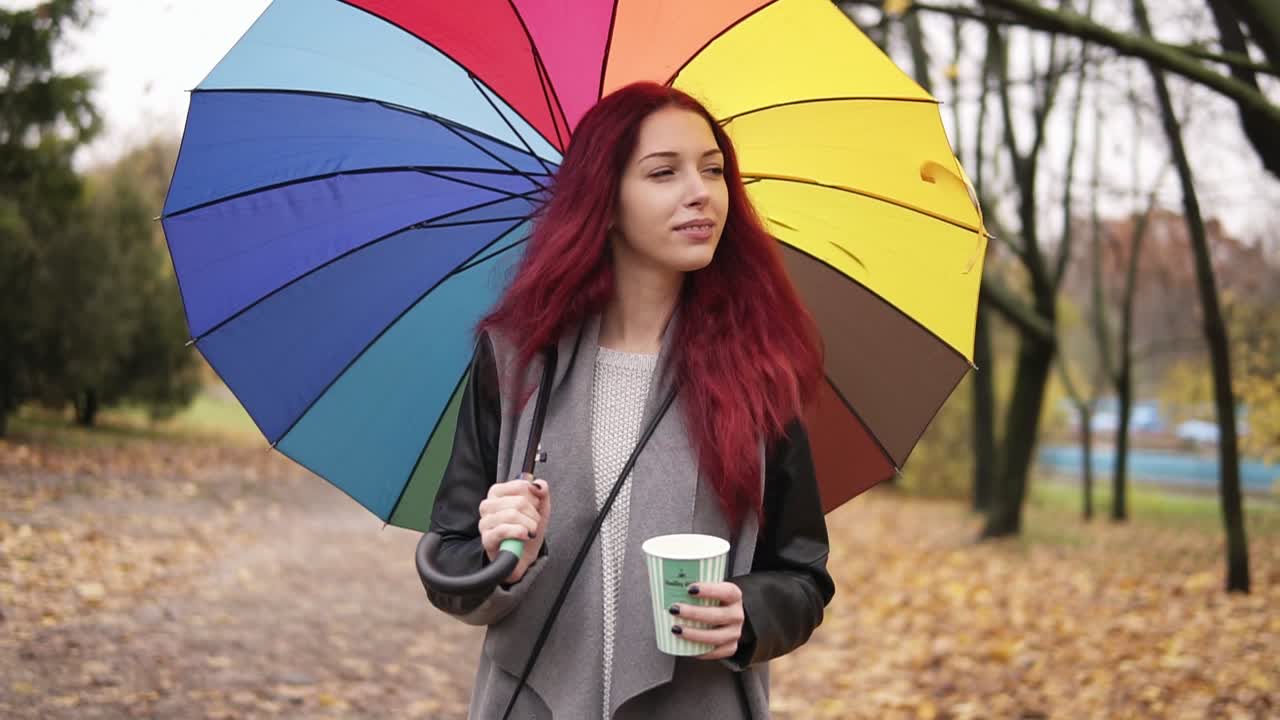 mujer joven de cabello rojo caminando en el parque de otoño y bebiendo café de una taza de papel mientras sostiene un paraguas de colores. niña con abrigo cálido disfrutando del clima de otoño fresco con una taza de bebida caliente