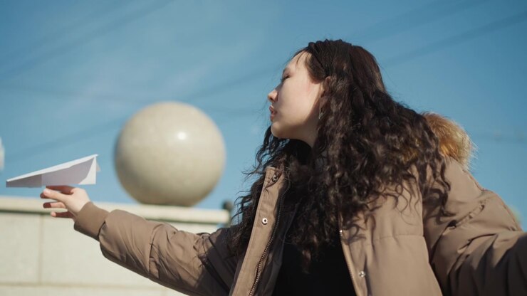 Una giovane donna si prepara a lanciare un aeroplanino di carta bianco sullo sfondo di una giornata soleggiata. La donna aggiusta la presa sull'aereo piegato mentre si trova vicino a strutture urbane e architetture sferiche.