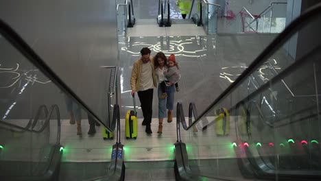 Familia-Feliz-Y-Joven-Con-Maleta-Amarilla-Subiendo-Por-La-Escalera-Mecánica-Desde-El-Aeropuerto