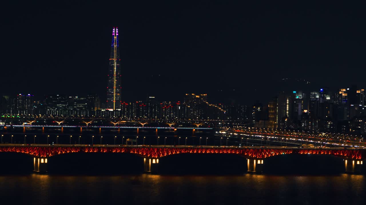 La vista aerea si sposta a destra lungo il ponte Seongsu arancione brillante sul fiume Han, con la Lotte World Tower e lo scintillante skyline notturno di Seoul che si estende in lontananza.