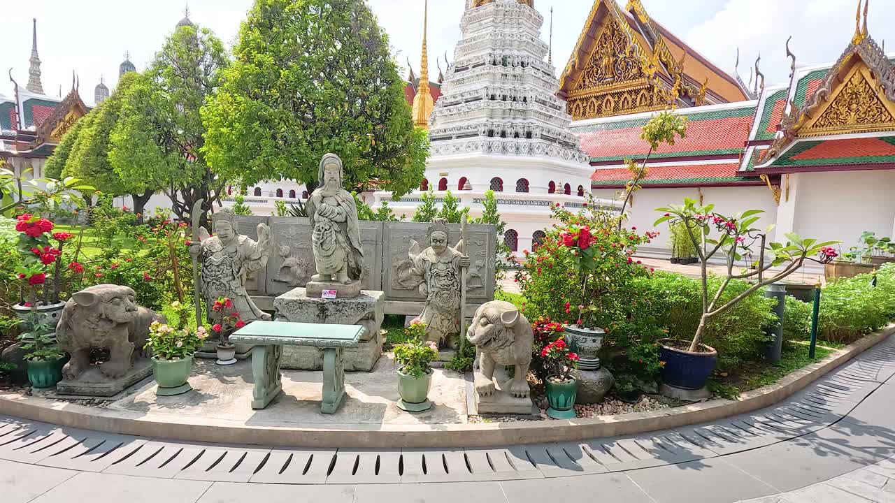 A tranquil garden scene at Wat Phra Kaew, featuring statues, lush greenery, and traditional architecture under bright daylight