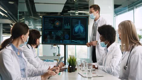 Multiethnic-doctors-team-wearing-facial-mask-discussing-covid-lung-disease-while-they-observing-digital-display-screen-in-hospital-office