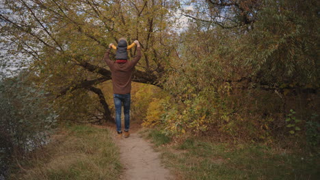 man is spending time with his child son at weekend walking together in park or forest picturesque autumn landscape toddler is sitting on shoulders of father rear view