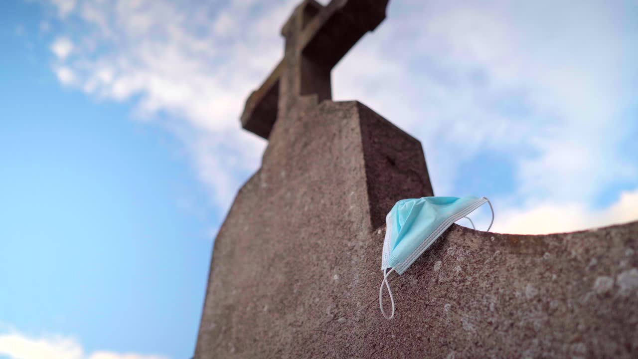 Death by Covid-19 pandemic, protective face mask discarded on a gravestone or tombstone with a religious cross in a graveyard or cemetery