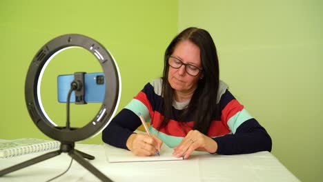 Adult-Woman-Wears-Eyeglasses-And-Write-Down-Notes-On-The-Desk-During-Online-Meeting