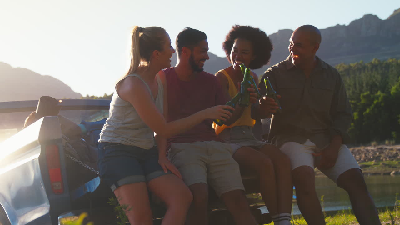 Friends With Backpacks Sitting On Tailgate Of Pick Up Truck On Road Trip By Lake Drinking Beer