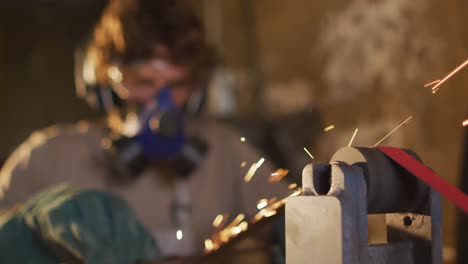 Caucasian male blacksmith wearing breathing mask and ear guards, forging metal tool in workshop