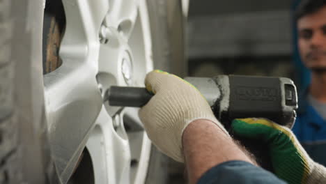 close-up of engineer in blue uniform using pneumatic tool to fix car wheel bolts in a garage, a colleague attentively observes, nodding his head, ensuring precision in vehicle maintenance