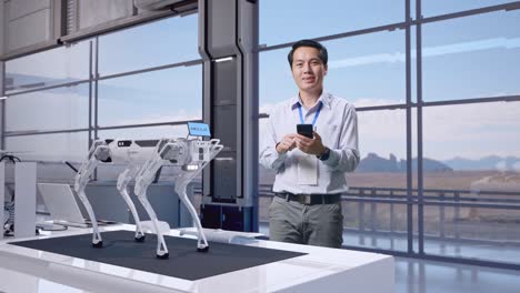 cuerpo lleno de un trabajador profesional masculino asiático de pie con su teléfono inteligente en una fábrica de alta tecnología, perro robot, está mirando a la cámara con una sonrisa