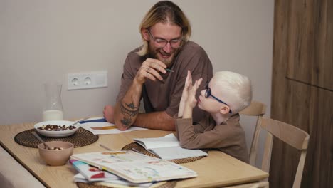 happy blond man with a beard and glasses helps his little albino son with white blond hair in blue glasses to count a math assignment using his fingers on his hand and do his homework while preparing for the next day at school in a modern kitchen at home